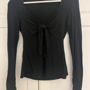 Svart långärmad topp med knyt och öppning fram - Säljer en svart långärmad topp med snygg knytning framtill. Toppen har en öppning fram  och är tillverkad i ett mjukt, stretchigt material. Perfekt att styla med jeans eller kjol för en trendig look.