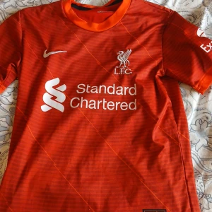 Liverpool FC matchtröja Nike - Säljer en röd Liverpool FC fotbollströja från Nike med Standard Chartered som sponsortryck. Tröjan är kortärmad och har vita detaljer samt klubbmärke på bröstet. Tillverkad i Dri-Fit material för bra andningsförmåga. Perfekt för fotbollsträning eller support.