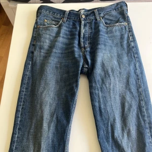 Blå jeans från Carhartt - Säljer ett par klassiska blå jeans från Carhartt med raka ben och fem fickor. Jeansen har en snygg tvätt och detaljer som läderpatch med logga bak samt metallknapp framtill. Perfekta för en avslappnad stil. en del slitage längst nere på benen men utöver de så är dom i bra skick