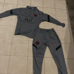 Grå Paris Saint-Germain fotbollströja med byxa - Säljer en grå långärmad fotbollströja från PSG med svarta detaljer och tillhörande byxa. Tröjan har klubbmärke och Nike-logga på bröstet samt texten 'ALL' och 'VISIT RWANDA'. Perfekt för träning eller uppvärmning.