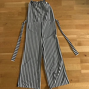 Svartvit randig byxdress/jumpsuit med knytband - Säljer en svartvit randig byxdress med vida ben och knytband i midjan. Står ingen storlek men skulle gissa på XS. Byxdressen har resår upptill och är ärmlös, vilket ger en avslappnad och trendig look. Går nästan till anklarna på mig som är 163 cm. Finns en liten defekt som syns på andra bilden, men knappt använd. Köpt utomlands! 