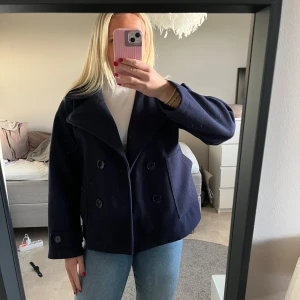 Marinblå kappa från design by si - Säljer en mörkblå kappa med dubbelknäppning och stora slag. Jackan har långa ärmar med knappdetalj vid ärmslut och två fickor framtill. Klassisk och stilren modell som passar till många tillfällen. Den är endast använd ett fåtal gånger och är i mycket fint skick! 