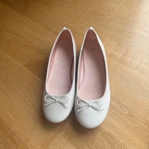 Vita ballerinaskor från H&M - Endast använda 1 gång, då de är för små för mig som har 37. Säljer ett par vita ballerinaskor från H&M med en söt rosett på tån. Skorna har en rund tå och en mjuk rosa innersula för extra komfort. Perfekta för en stilren och enkel look.