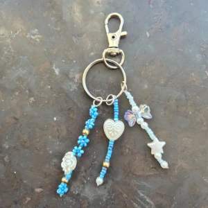 Nyckelring med pärlor och berlocker - Säljer en söt handgjord nyckelring med tre hängen av blå och vita pärlor, dekorerad med berlocker i form av hjärta, blomma, stjärna och fjäril. Perfekt som accessoar till väskan eller nycklarna.