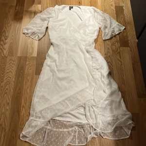 Vit omlottklänning med volang från Vero Moda - Säljer en vit omlottklänning från Vero Moda med volangdetaljer och knyt i midjan. Klänningen har halvtransparenta, korta volangärmar och subtilt prickigt mönster i tyget. Perfekt för sommarens tillfällen! Den är helt ny, har aldrig använts.