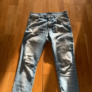 Ljusblå slitna jeansbyxor från nudie jeans - Ljusblå jeansbyxor med slitningar och hål på båda benen. Klassisk femficksmodell med orangea sömmar och knappgylf. Jeansen har en rakare och tapered passform och är tillverkade i denim. Perfekta för en avslappnad och trendig look. lite slitet på bakfickan som ni kan se men tycker det ser fett ut höger ficka har en liten defekt även