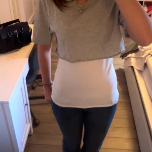 Grå croppad t-shirt - Säljer en grå croppad t-shirt i bomull som bärs ovanpå en vit tight linnetopp. Perfekt för en avslappnad och trendig look. T-shirten har korta ärmar.
