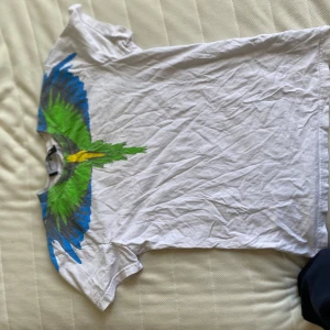 Vit t-shirt med papegojtryck från Marcelo Burlon - Säljer en vit t-shirt från Marcelo Burlon med ett stort färgglatt papegojtryck över axlarna i grönt, blått och gult. T-shirten har normal passform och är kortärmad. Perfekt för dig som vill sticka ut med en unik design.