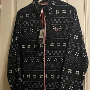 Ny fleecejacka från Swedemount! 42 - Säljer en svart fleecejacka från Swedemount med ett vitt mönster och rosa dragkedja. Jackan har långa ärmar och en hög krage, perfekt för kyliga dagar. Den är både stilren och funktionell. Prislappar kvar 