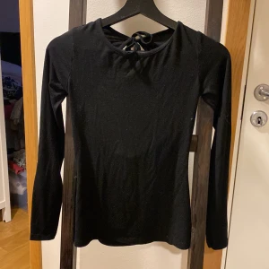 Svart långärmad topp med knyt och öppen rygg - Säljer en svart långärmad topp med snygg knytning i nacken och öppen rygg. Toppen är figurnära och passar perfekt till många olika outfits. Mjuk och stretchig bomullsblandning.