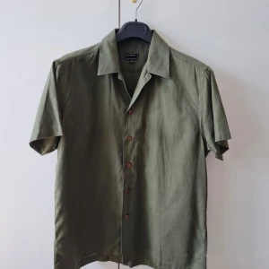 Kortärmad herrskjorta från Eastwest strl M - Snygg olivgrön skjorta från Eastwest i storlek M. Skjortan har korta ärmar, klassisk krage och knäppning framtill med bruna knappar. Perfekt för varma dagar och enkel att matcha med jeans eller shorts. Tillverkad i bomull. Bröstmått 114cm. Storlek M. Kan hämtas i Vallentuna.