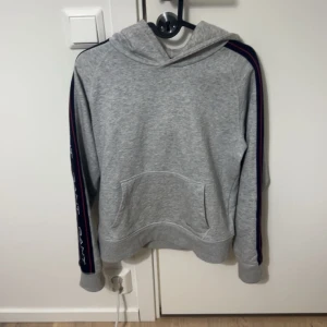 Grå hoodie från GANT - Säljer en stilren grå hoodie från GANT med mörkblå och röda ränder längs ärmarna. Tröjan har en klassisk känguruficka och en bekväm huva. Perfekt för en avslappnad look.