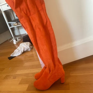 Orange stövlar med klack - Snygga orange stövlar i mocka med hög klack och över knäet-design. Perfekta för att göra ett statement med din outfit. De har en rund tå och en stabil klack för extra komfort. Endast använd en gång inomhus på halloweenfest 