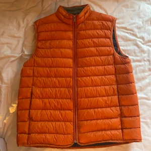 Orange dunväst från Hampton Republic - Säljer en snygg orange dunväst från Hampton Republic. Västen har en dragkedja framtill och är quiltad för extra värme. Insidan är i en kontrasterande mörkblå färg med gröna detaljer. Perfekt för kyliga dagar!