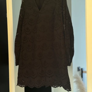 Svart klänning med spetsdetaljer - Elegant svart klänning med v-ringning och vackra spetsdetaljer. Klänningen har långa ärmar och ett genombrutet mönster som ger en stilfull look. Perfekt för en chic och sofistikerad stil.