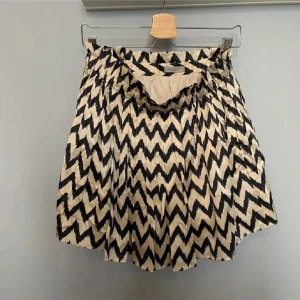 Mönstrad kjol från Hunkydory - Snygg kortkjol från Hunkydory med ett coolt zigzag-mönster i svart och beige. Kjolen är tillverkad i silke med polyesterfoder, vilket ger en lyxig känsla. Perfekt för att ge din outfit en trendig touch! Köpt över 2000 kr