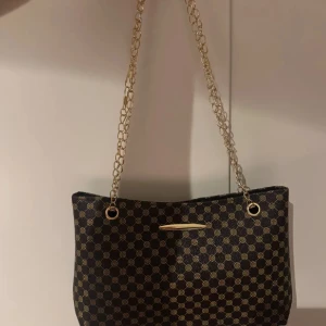 Mönstrad axelväska med kedja - Snygg axelväska med ett elegant mönster i svart och guld. Väskan har en stilren kedja som axelrem och en diskret metallplatta på framsidan. Perfekt för att ge din outfit en lyxig touch.