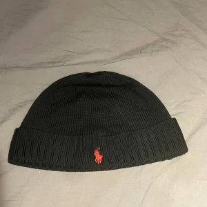 Svart mössa från Polo Ralph Lauren - Snygg svart mössa från Polo Ralph Lauren med röd broderad logotyp framtill. Mössan är ribbad och tillverkad i ett mjukt material som passar perfekt för kyligare dagar.