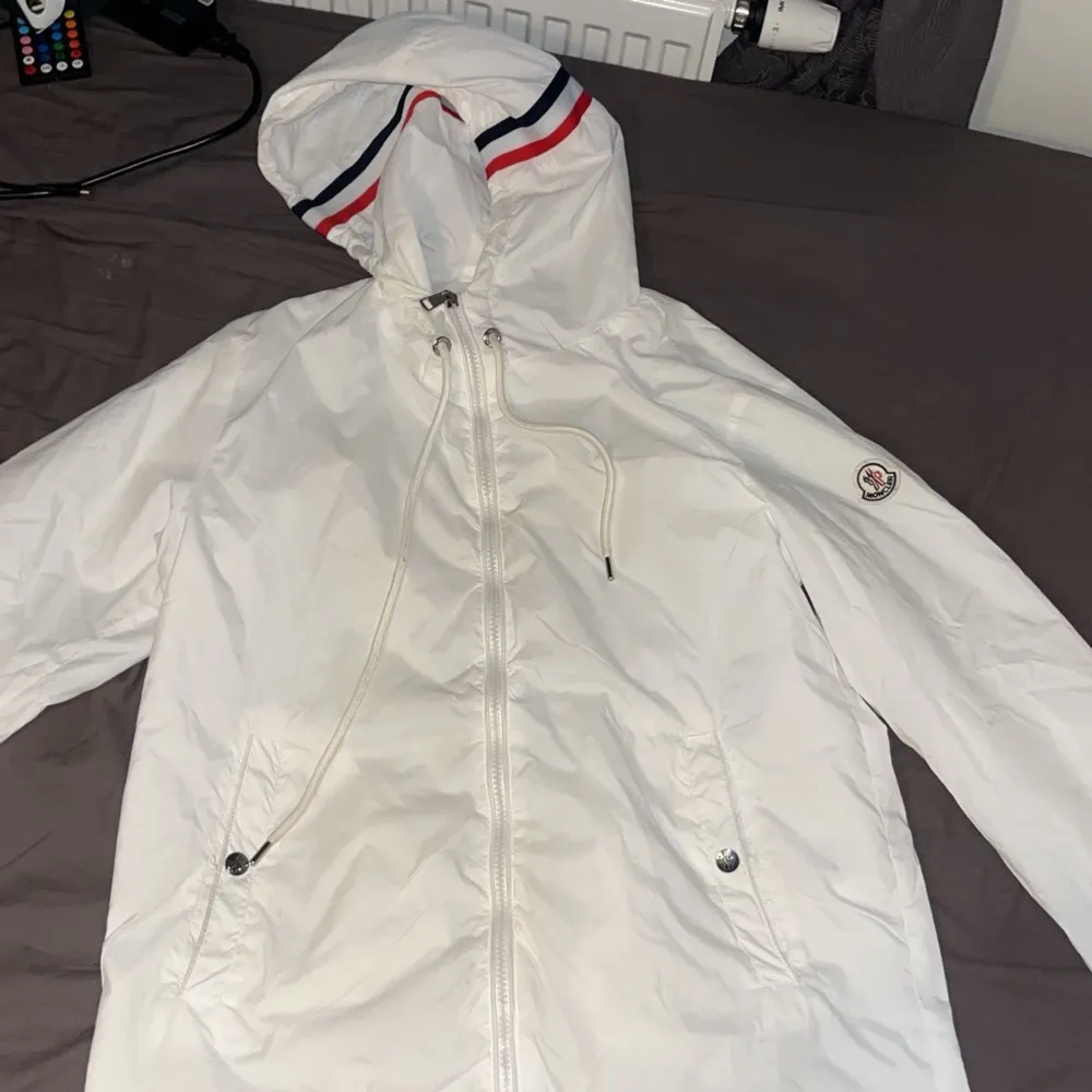 Säljer en stilren vit vindjacka från Moncler med dragkedja och justerbar huva. Jackan har Moncler-logga på ärmen och röd-blå randdetalj på huvan. Perfekt för vår och sommar.. Takit.