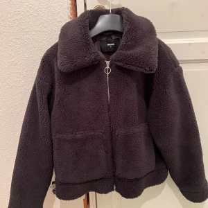 Svart teddyjacka från BikBok - Mysig svart teddyjacka från BikBok med dragkedja framtill. Jackan har en stor krage och två framfickor, perfekt för kyliga dagar. Den är både stilren och bekväm, ett måste i garderoben! Helt oanvänd.