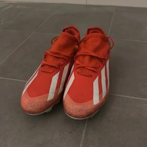Röda fotbollsskor från Adidas - Snygga röda fotbollsskor från Adidas med vita ränder och snörning. Perfekta för fotbollsplanen med en stilren design och bra passform.