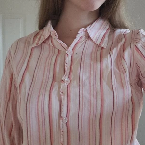 Randig skjorta i rosa och vit - Säljer en stilren randig skjorta i rosa och vit med knappar framtill. Skjortan har en klassisk krage och långa ärmar, perfekt för en avslappnad men ändå uppklädd look.