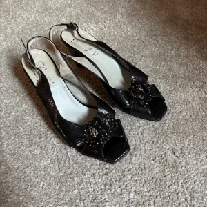 Svarta klackskor med blommig detalj - Snygga svarta klackskor i äkta läder med öppen tå och en elegant blommig detalj med pärlor framtill. Tillverkade i Italien, dessa skor har en stilren design med en bekväm klack och justerbar rem runt hälen.