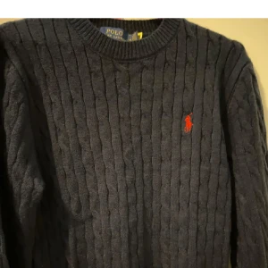 Mörkblå kabelstickad tröja från Ralph Lauren - Säljer en stilren mörkblå kabelstickad tröja från Ralph Lauren med det klassiska röda logotypbroderiet på bröstet. Tröjan har en rund halsringning och är perfekt för kyligare dagar. Passar både till jeans och chinos för en avslappnad look.