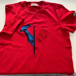 Röd t-shirt från Moncler - Snygg röd t-shirt från Moncler med en broderad tupp i blått och rött på framsidan. T-shirten har korta ärmar och Monclers logga på ärmen. Använd ett fåtal gånger.