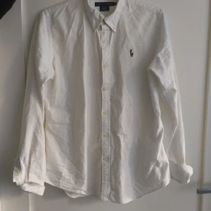 Vit skjorta från Ralph Lauren - Säljer en stilren vit skjorta från Ralph Lauren i slim fit. Skjortan är tillverkad i 100% bomull och har långa ärmar. Den har en klassisk krage och knappar framtill. På bröstet finns det ikoniska broderade logotypen med en polospelare.Använd endas 1 gång. Öppen för prisförslag 