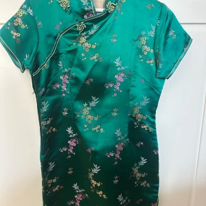 Grön sidenklänning med blommönster - Elegant grön klänning i siden med vackert blommönster i rosa, gult och vitt. Klänningen har en traditionell design med korta ärmar och en unik krage. Perfekt för speciella tillfällen.