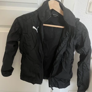 Svart vindjacka från Puma - Säljer en svart vindjacka från Puma med dragkedja och huva. Jackan har en stilren design med Puma-loggan på bröstet. Perfekt för blåsiga dagar och lätt att packa med sig. Passar både för träning och vardag.