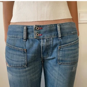 Blå jeans med dubbla knappar - Snygga blå jeans från Diesel med dubbla knappar i midjan och coola fickdetaljer. De har en straight passform och är perfekta för en avslappnad stil. Märkesdetaljer syns på bakfickan och bältesslingan. Innerbenslängd 77cm, midja 90 cm (runt)