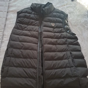 Svart dunväst från Emporio Armani - Snygg svart dunväst från Emporio Armani med dragkedja och ett stilrent märke på bröstet. Perfekt för lager-på-lager under kyligare dagar. Västen har en hög krage för extra värme och komfort. 1:1a