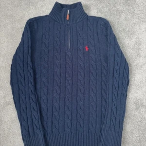 Mörkblå stickad tröja med dragkedja från Ralph Lauren - Säljer en snygg mörkblå stickad tröja från Ralph Lauren med dragkedja och röd broderad logga på bröstet. Tröjan har en kabelstickad design och hög krage, perfekt för kyliga dagar.