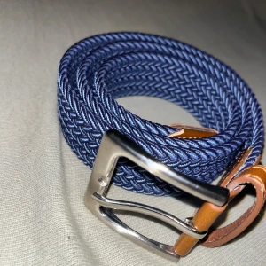 Blått flätat bälte med bruna detaljer - Snyggt blått flätat bälte med bruna läderdetaljer och en stilren silverfärgad spänne. Perfekt för att ge en casual look en extra touch. Passar både till jeans och chinos.
