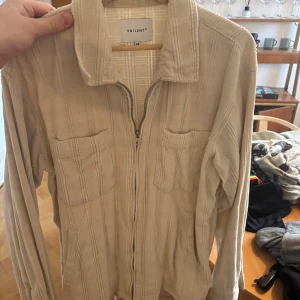 Beige overshirt från Vailent i Manchester material - Säljer en stilren beige overshirt från Vailent med dragkedja framtill. Skjortan har två bröstfickor och ett diskret randigt mönster. Perfekt för lager-på-lager-stil.