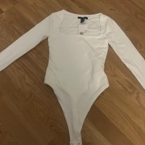 Vit långärmad bodysuit från Forever 21 - Säljer en stilren vit långärmad bodysuit från Forever 21. Den har en fyrkantig urringning och är perfekt för att skapa en snygg och enkel look. Materialet är mjukt och stretchigt för en bekväm passform. Perfekt att matcha med jeans eller kjol!