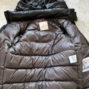 Svart dunjacka från Moncler - Säljer en stilren svart dunjacka från Moncler. Jackan har en glansig yta och är perfekt för kyligare väder. Den har en dragkedja framtill och en ikonisk Moncler-logga på insidan. Perfekt för dig som vill hålla stilen i vinter.