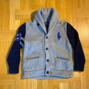 Polo Palph Laurend 🇸🇪🌟 - Säljer en snygg grå och marinblå kofta från Ralph Lauren med knappar framtill och en broderad logga på bröstet. Koftan har långa ärmar och en stilren krage. Perfekt för kyligare dagar!