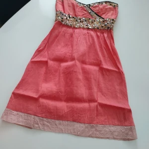 Rosa klänning från Valerie i SIDEN. - Snygg rosa klänning från Valerie i äkta SIDEN av högsta kvalitet med glittrande paljettdetaljer runt bysten. Klänningen är axelbandslös och har en elegant design med en lätt glansig finish. Perfekt för festliga tillfällen!