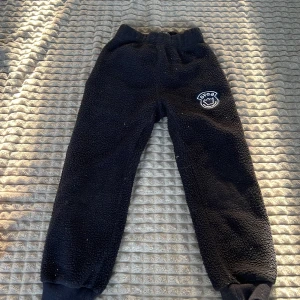 Svarta fleecebyxor med broderad detalj - Mysiga svarta fleecebyxor med elastisk midja och ribbade muddar vid bensluten. Byxorna har en broderad detalj på ena benet som ger en unik touch. Perfekta för kalla dagar.