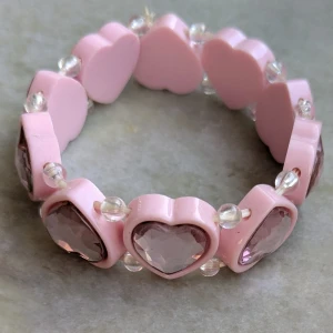 Rosa armband med hjärtformade detaljer - Charmigt rosa armband med hjärtformade pärlor och glittriga stenar. Armbandet har genomskinliga pärlor mellan varje hjärta, vilket ger en söt och lekfull look. Passar barn