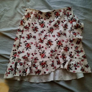 Blommig omlottkjol med volang - Söt blommig kjol från NLY One i vitt med röda blommor. Kjolen har en volangdetalj längs nederkanten. Perfekt för sommarens alla tillfällen!