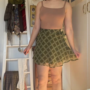 Grön rutig minikjol - Köpt secondhand men aldrig använd. Säljer en söt grön rutig kortkjol med omlott-design och knytning på sidan. Perfekt för en avslappnad och stilren look. Kjolen har en lätt och luftig känsla, vilket gör den idealisk för varmare dagar.