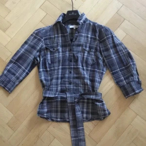 Rutig skjorta med bälte från Cue - Snygg rutig skjorta från Cue i grå och blå toner. Skjortan har trekvartsärmar och ett matchande bälte i midjan för en stilren look. Perfekt för en avslappnad men ändå chic stil. 