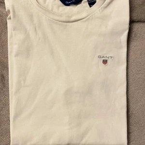 Vit t-shirt från GANT - Säljer en stilren vit t-shirt från GANT med broderad logga på bröstet. T-shirten är kortärmad och tillverkad i mjuk bomull. Perfekt för en avslappnad look. Passar barn i åldern 11-12 år.