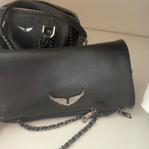 Svart zadig väska - Snygg svart handväska med kedjedetalj och ett dekorativt emblem i form av vingar. Väskan har en dragkedja och är perfekt för att bära dina nödvändigheter med stil. Den har en elegant och modern design som passar till många olika tillfällen.