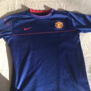 Blå Manchester United fotbollströja från Nike - Säljer en blå Manchester United fotbollströja från Nike med korta ärmar. Tröjan har röda detaljer och klubbens emblem på bröstet. Perfekt för fans som vill visa sitt stöd. Tillverkad i Thailand med Nike Fit Dry-teknologi.
