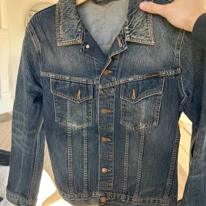 Jeansjacka från Nudie Jeans - Snygg jeansjacka från Nudie Jeans i klassisk blå denim. Jackan har knappar framtill och två bröstfickor med lock. Perfekt för en avslappnad stil.
